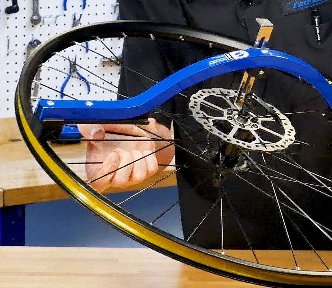 Hand squeezing two spokes of the same side near foot of wheel alignment gauge resting on rim